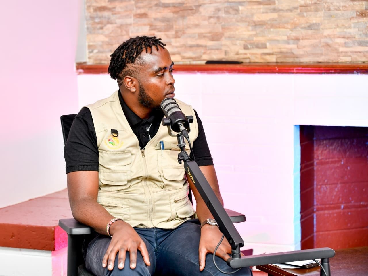 Denzel Maina in profile at a podcast microphone, wearing a black shirt and beige vest