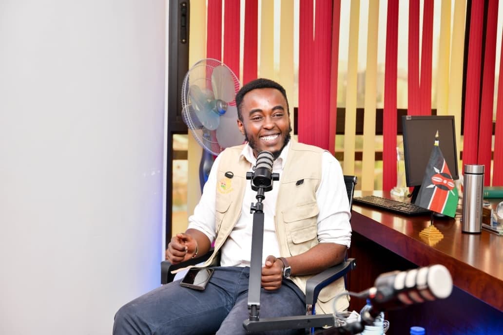 Wide shot of Denzel Maina at a microphone beside an office desk with a fan and a Kenyan flag